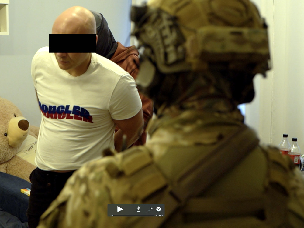 Mężczyzna w białej koszulce z logo stoi z rękami związanymi z tyłu, a w tle widoczny jest żołnierz w mundurze.