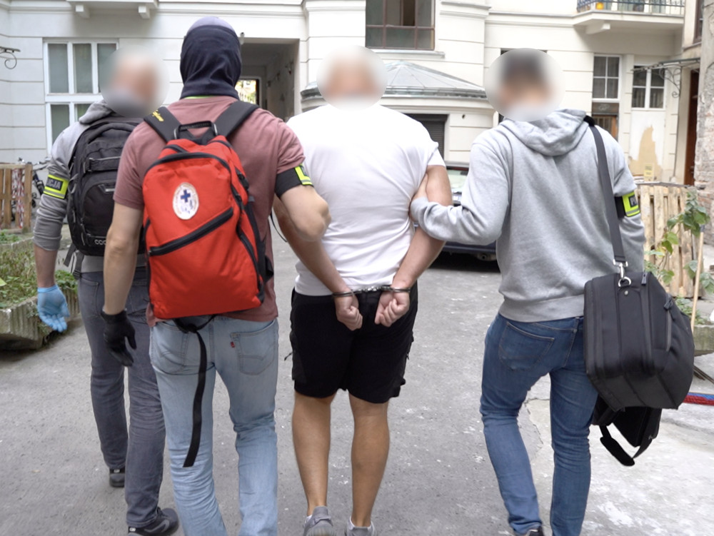 Mężczyzna w kajdankach prowadzony przez dwóch funkcjonariuszy w nieutwardzonej uliczce między budynkami.