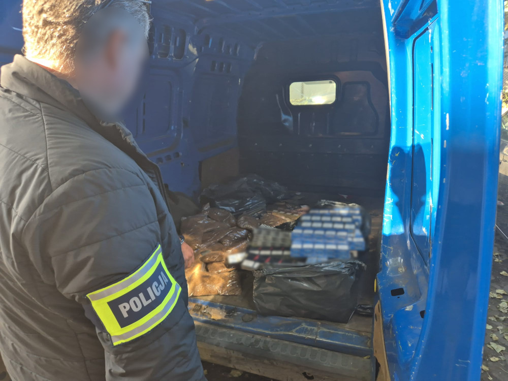 Policjant w mundurze sprawdzający zawartość niebieskiego furgonu, w którym znajdują się torby z nieznanym ładunkiem.