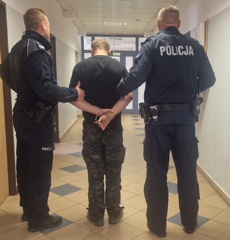 Mężczyzna w kajdankach prowadzony przez dwóch policjantów w korytarzu budynku.