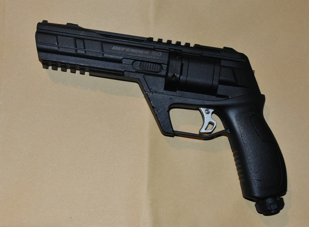 Czarny pistolet typu Defender 50 leży na beżowym tle, z widocznymi detalami konstrukcyjnymi i uchwytem.
