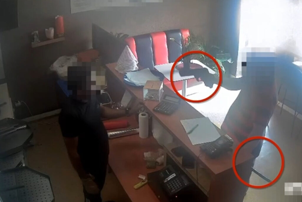 Mężczyzna w czarnej koszulce stoi przy ladzie, podczas gdy inny mężczyzna w pasiastym ubraniu trzyma pistolet.