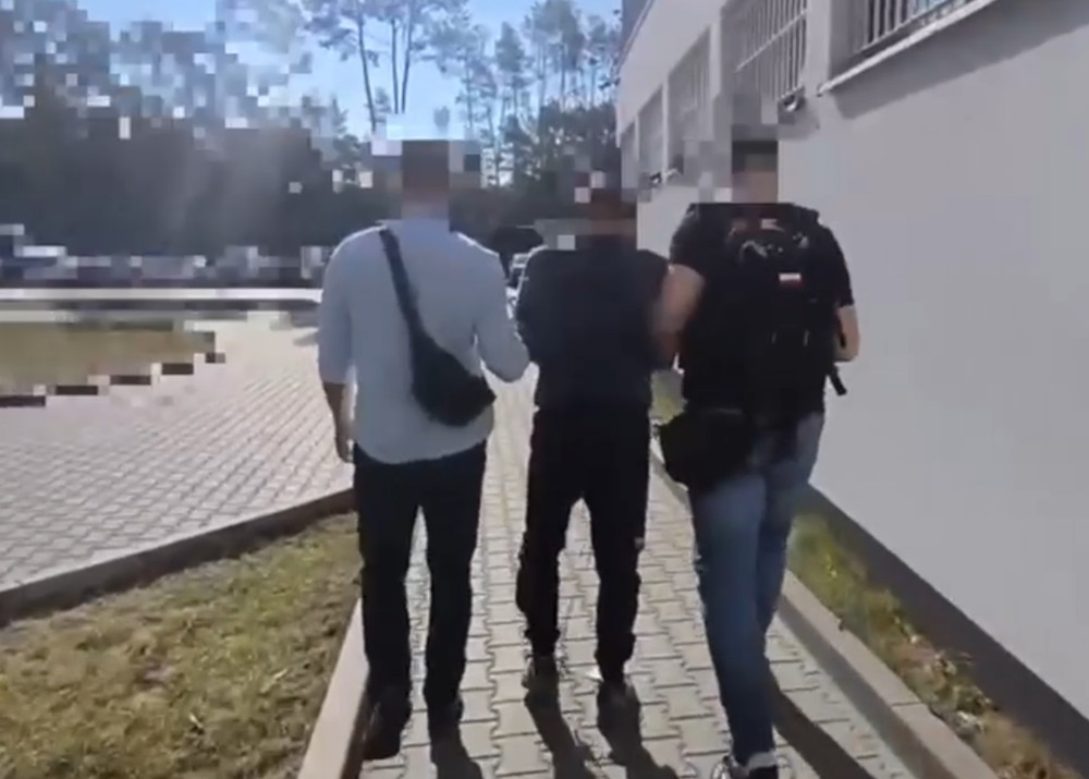 Trzech mężczyzn idzie chodnikiem wzdłuż budynku, jeden z nich jest prowadzony przez dwóch pozostałych.