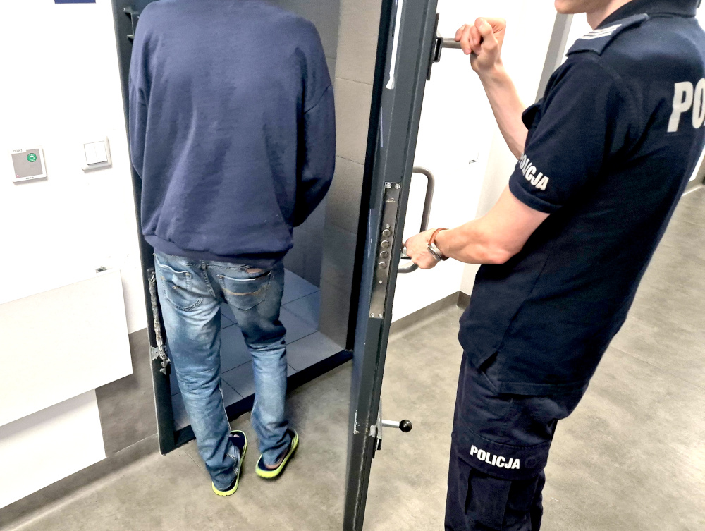 Mężczyzna w dżinsach i bluzie stoi tyłem przed otwartymi drzwiami celi, a policjant zamyka drzwi.