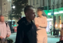 Mężczyzna w czarnej bluzie i krótkich spodenkach spaceruje po ulicy w nocy, obok grupy przechodniów.