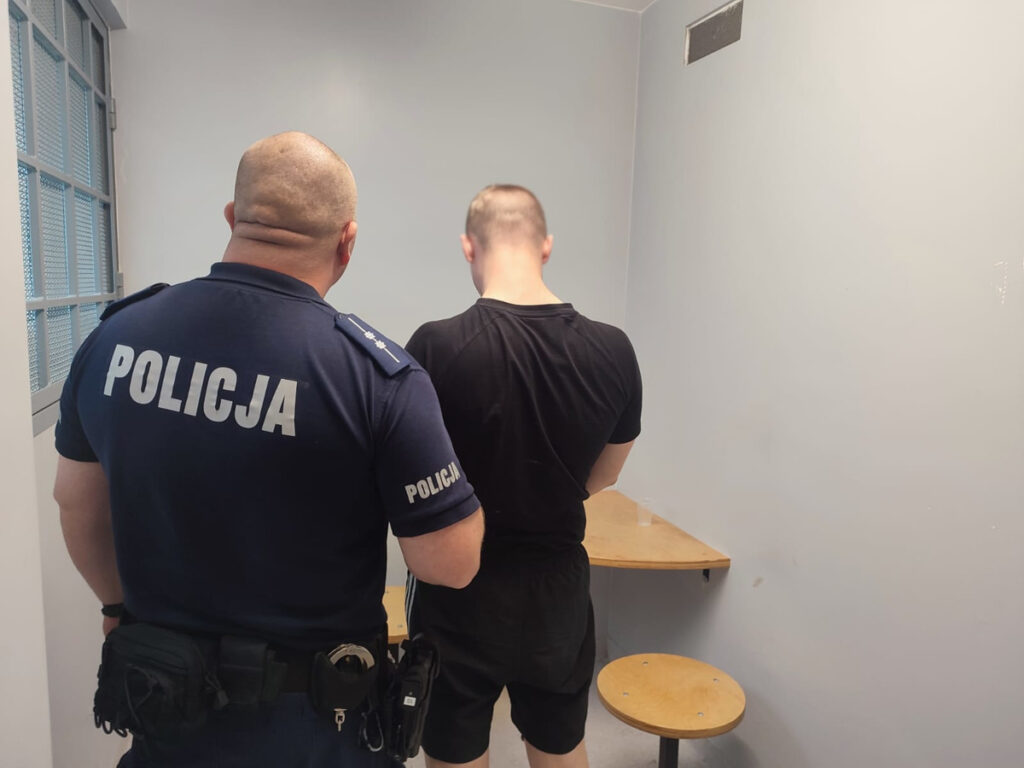 Policjant w mundurze obserwuje mężczyznę w czarnym T-shircie stojącego w pustym pomieszczeniu.