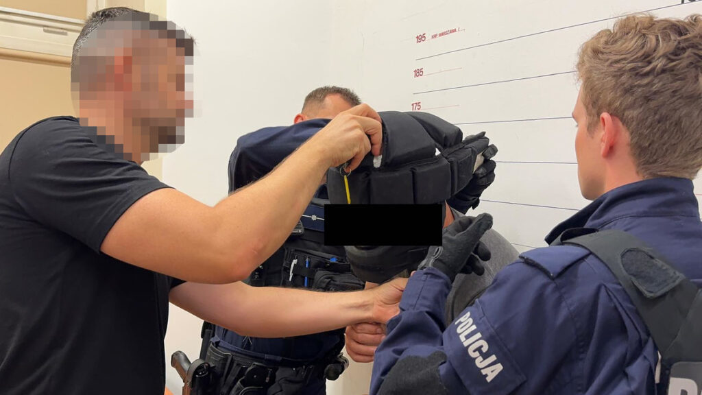 Mężczyzna w czarnym t-shircie zakłada hełm na głowę osoby w szarym ubraniu, w otoczeniu policjantów.