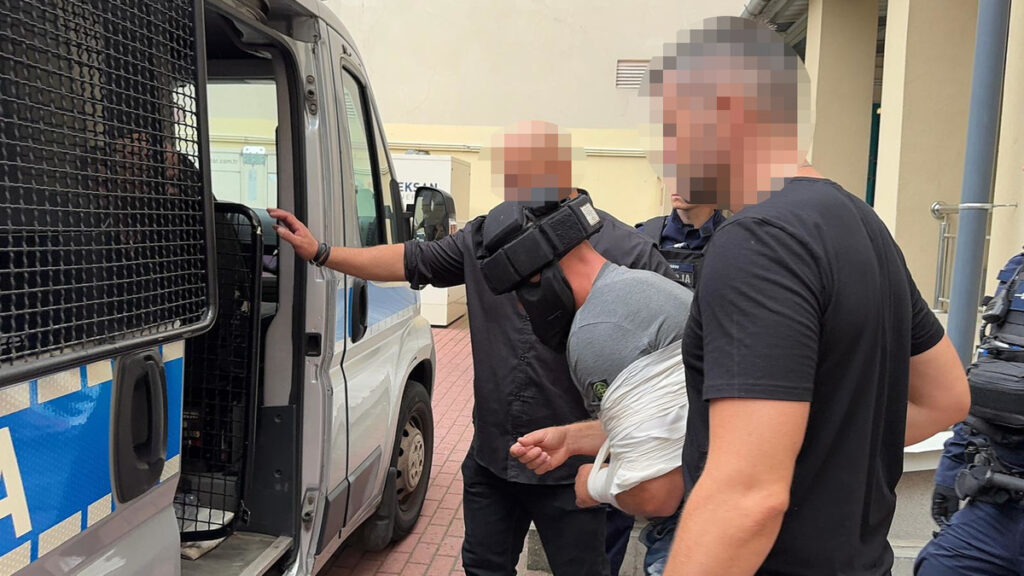 Mężczyzna w kajdankach wprowadzany do policyjnego furgonu przez funkcjonariuszy.