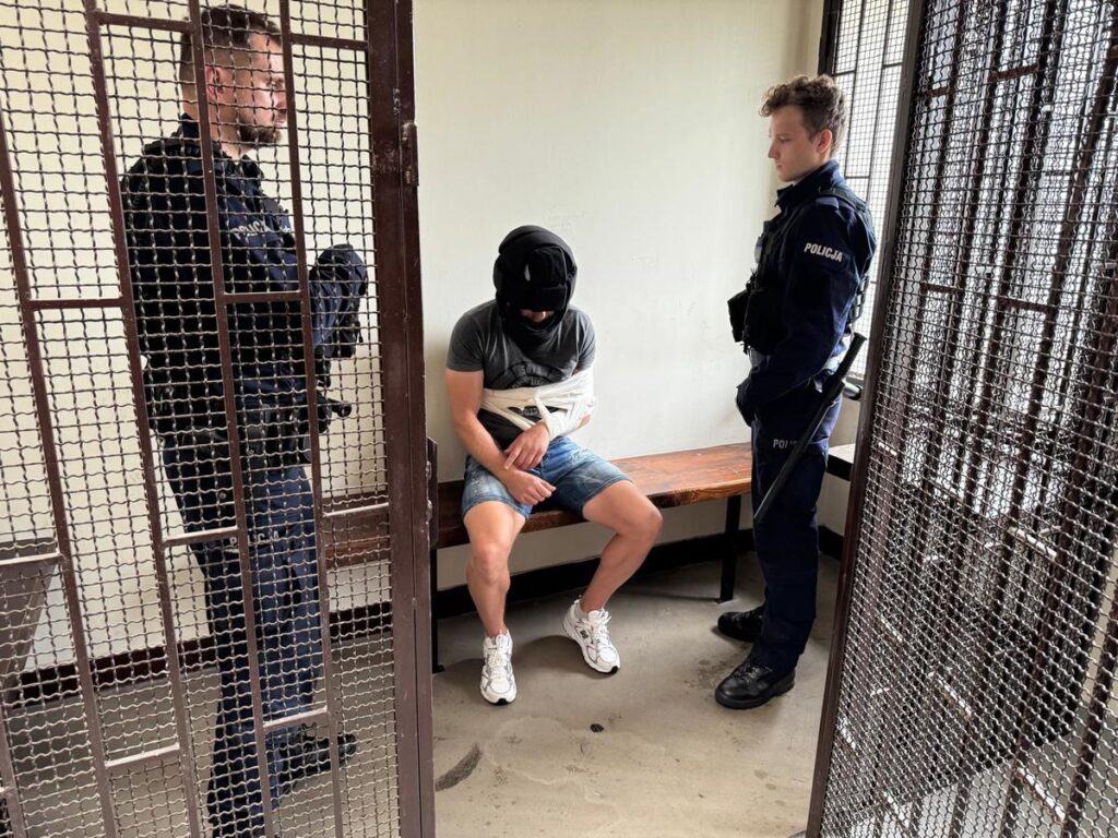 Mężczyzna w szortach i z zasłoniętą twarzą siedzi na ławce, podczas gdy dwóch policjantów stoi obok w zamkniętym pomieszczeniu.