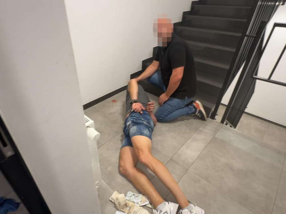 Mężczyzna w ciemnej koszulce pomaga leżącemu na schodach mężczyźnie w szortach, otoczeni przez szare ściany.