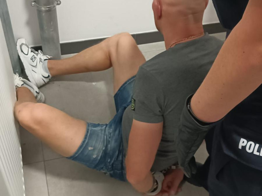 Mężczyzna w krótkich szortach siedzi na podłodze z rękami związanymi kajdankami, obok policjant w mundurze.