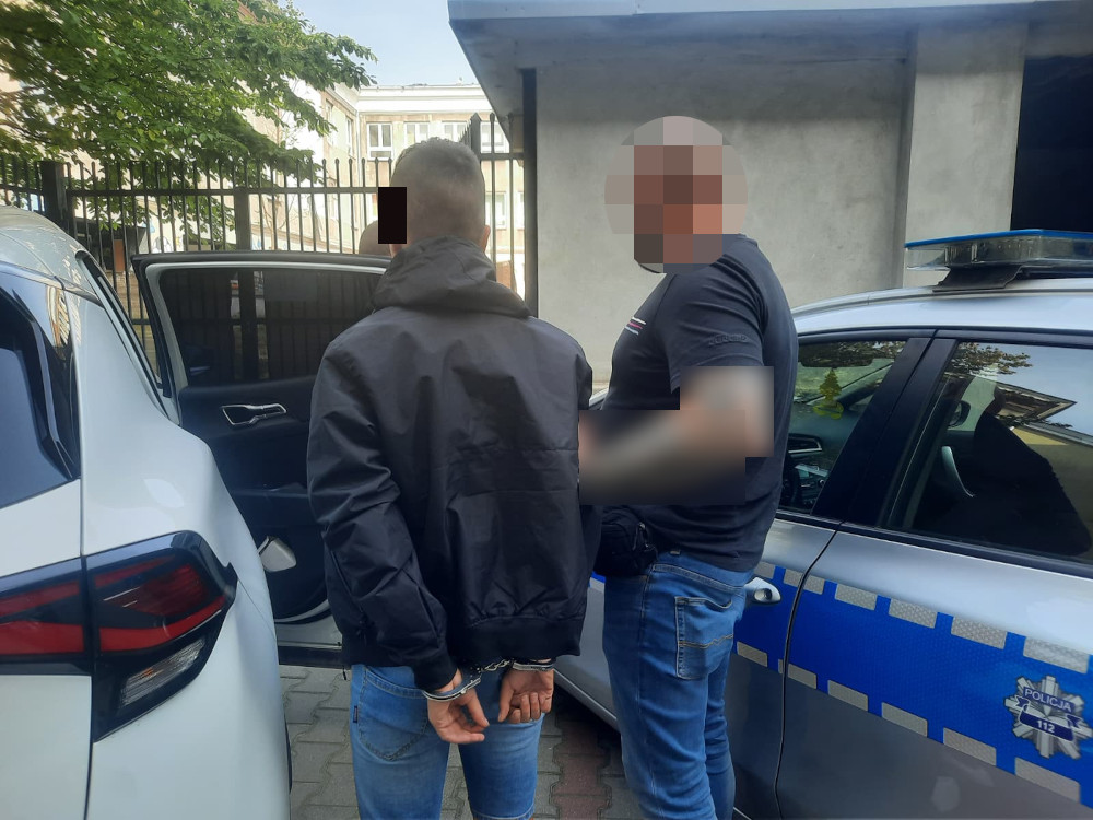 Mężczyzna w kajdankach stoi obok policyjnego samochodu, podczas gdy funkcjonariusz rozmawia z nim.