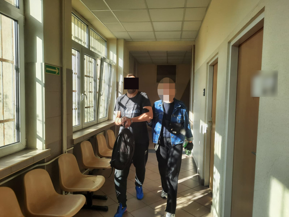 Dwaj młodzi mężczyźni idą korytarzem, trzymając się za ręce, w tle okna wpuszczające naturalne światło.