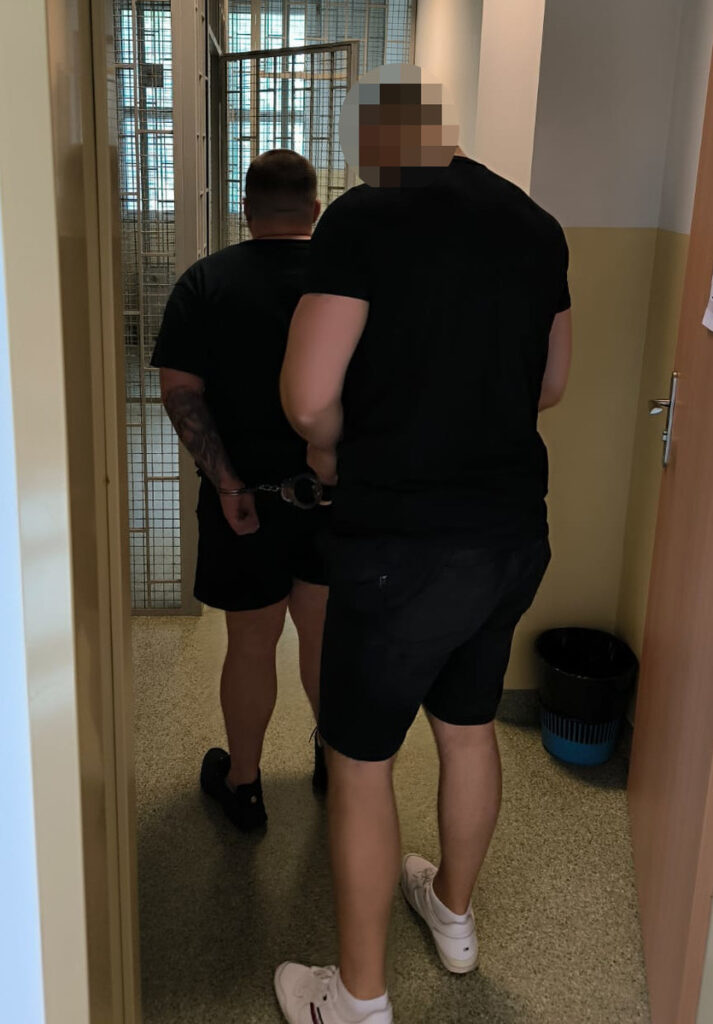 Mężczyzna w kajdankach prowadzony przez funkcjonariusza w korytarzu budynku policyjnego.