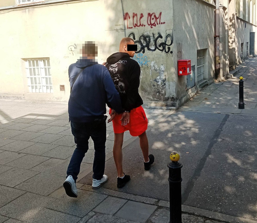 Mężczyzna w krótkich szortach prowadzony przez funkcjonariusza policji na ulicy z graffiti na ścianie.