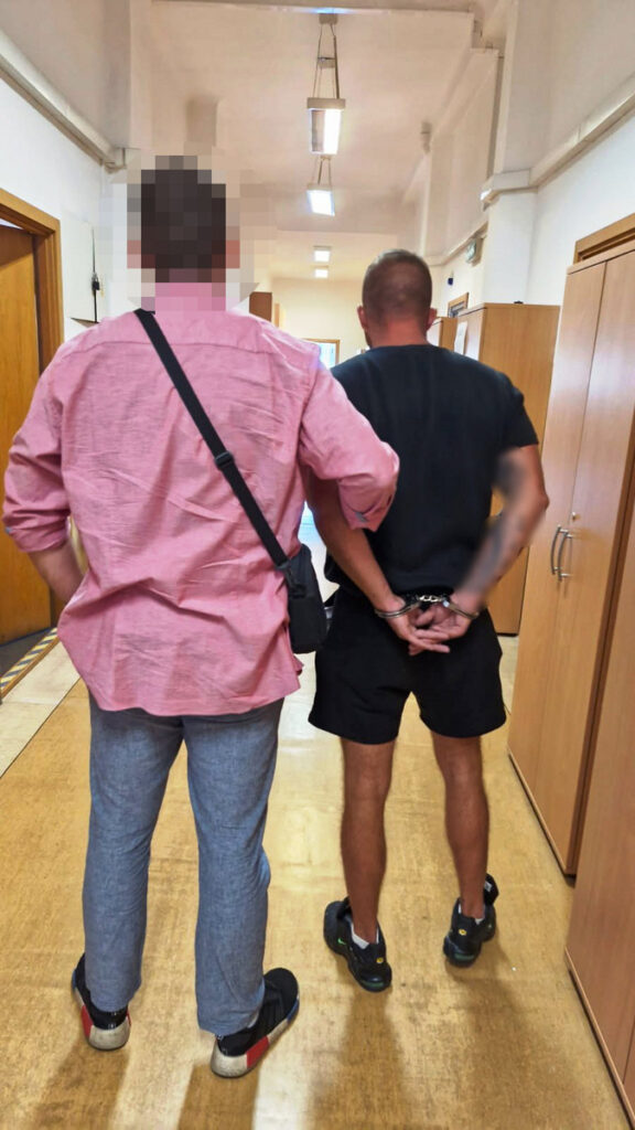 Mężczyzna w różowej koszuli prowadzi osobę w czarnej koszulce z rękami związanymi na plecach przez korytarz.
