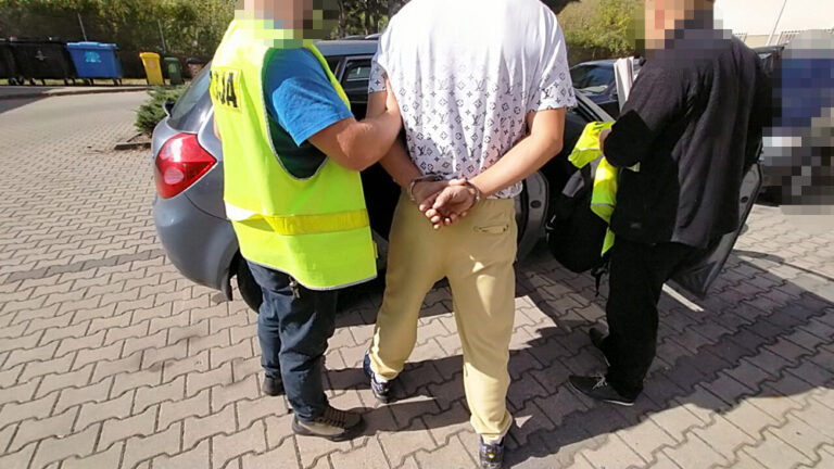 Mężczyzna w białej koszulce z logo, z rękami związanymi z tyłu, stoi obok samochodu, podczas interwencji służb.