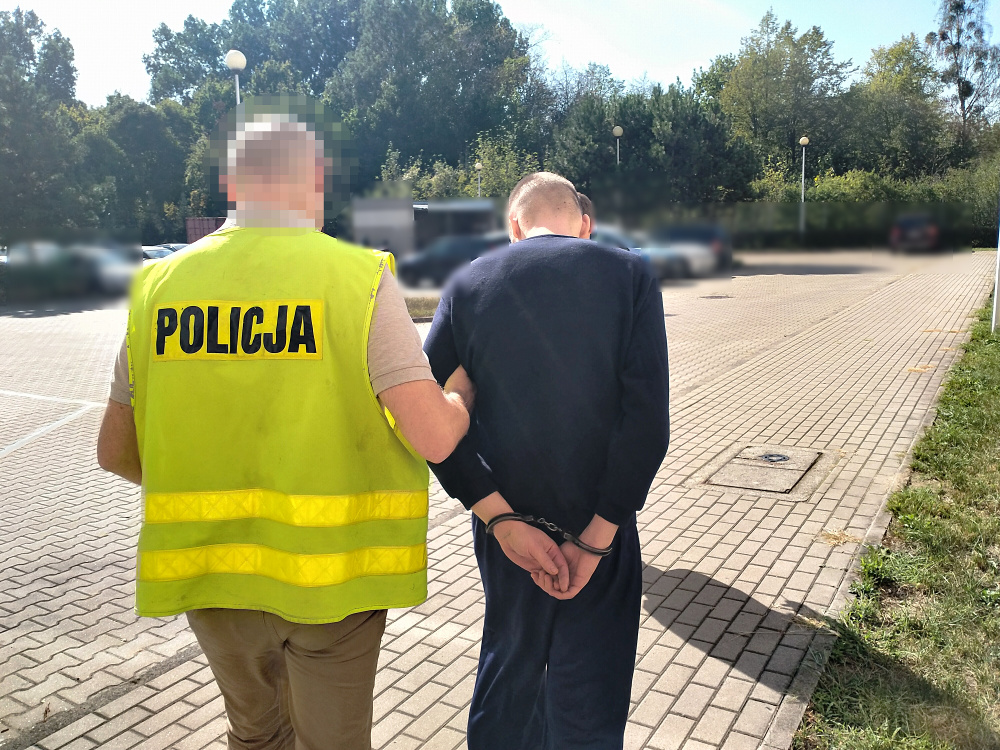 Mężczyzna w kajdankach prowadzony przez policjanta w kamizelce na parkingu otoczonym drzewami.