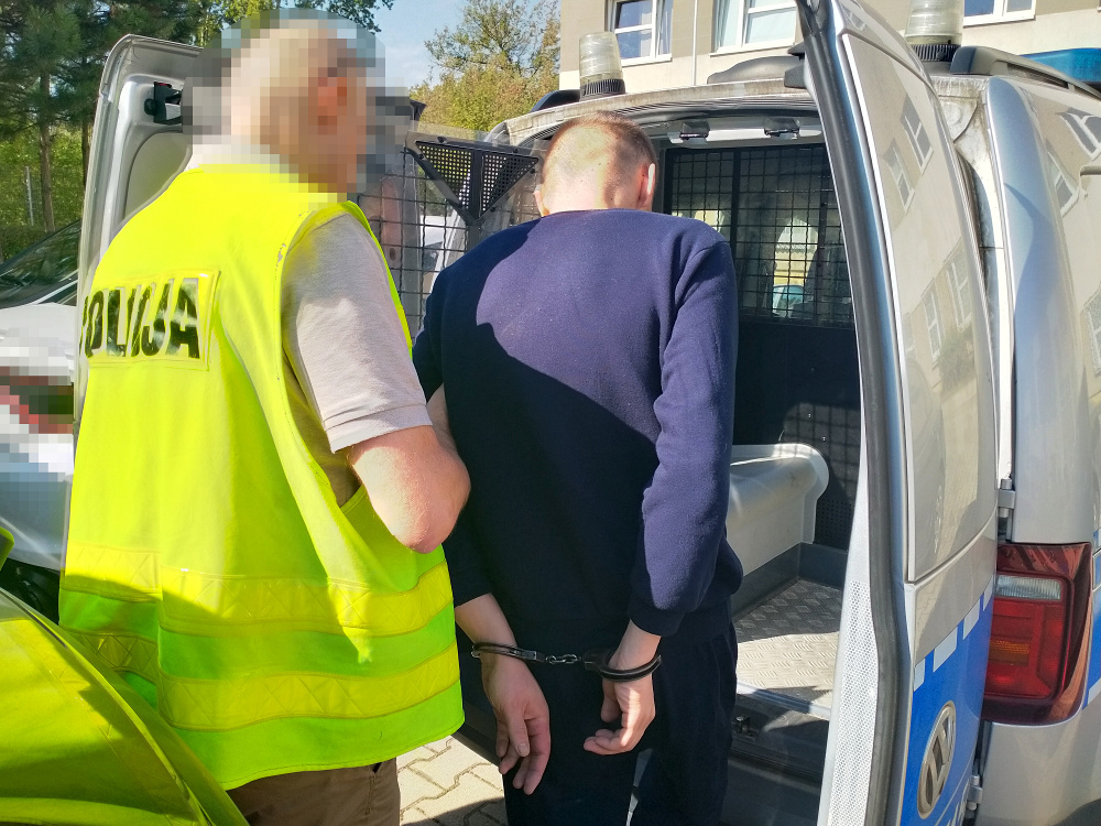 Mężczyzna w kajdankach wchodzi do policyjnego auta, obok stoi funkcjonariusz w kamizelce odblaskowej.