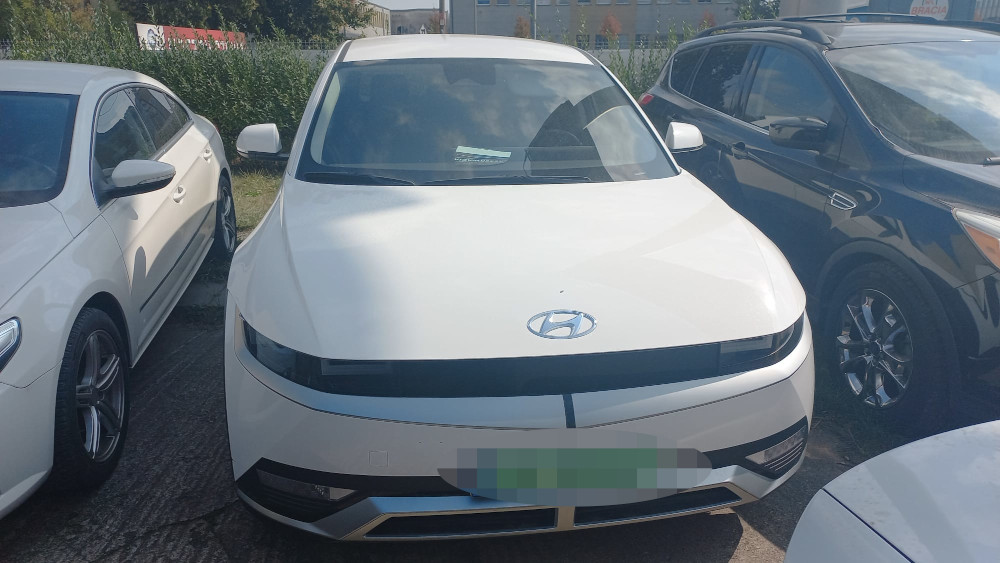Biały samochód osobowy zaparkowany pomiędzy dwoma innymi pojazdami na parkingu, otoczony zielenią.