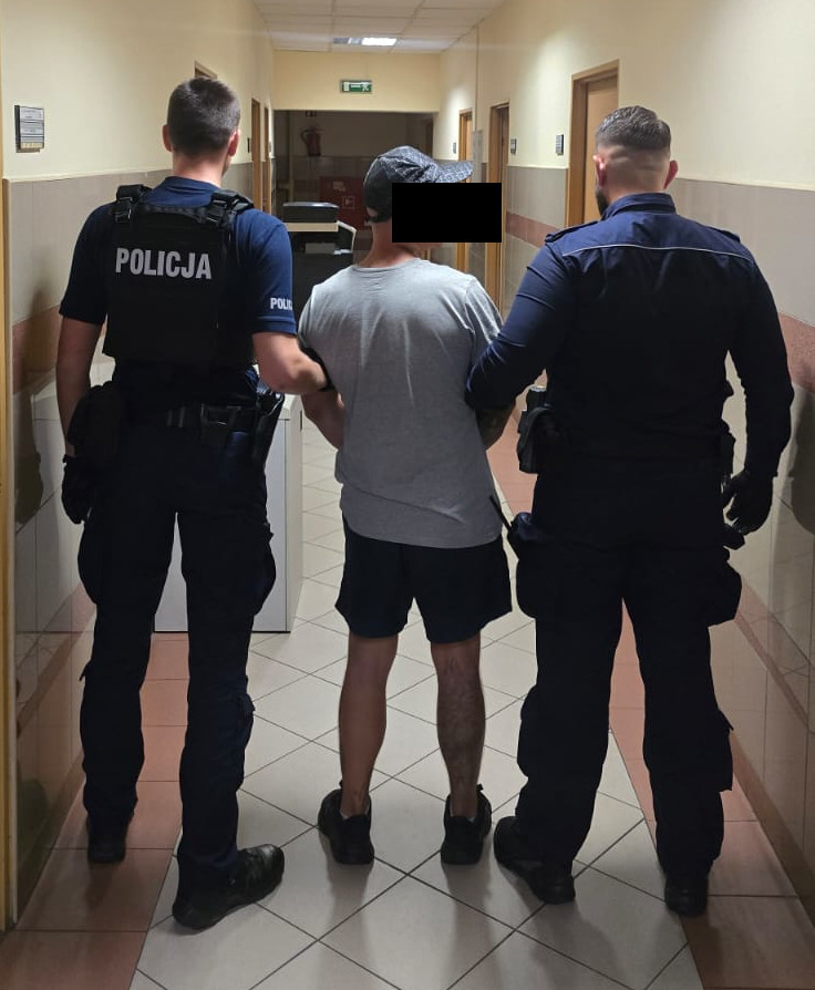 Mężczyzna prowadzony przez dwóch policjantów w korytarzu budynku.
