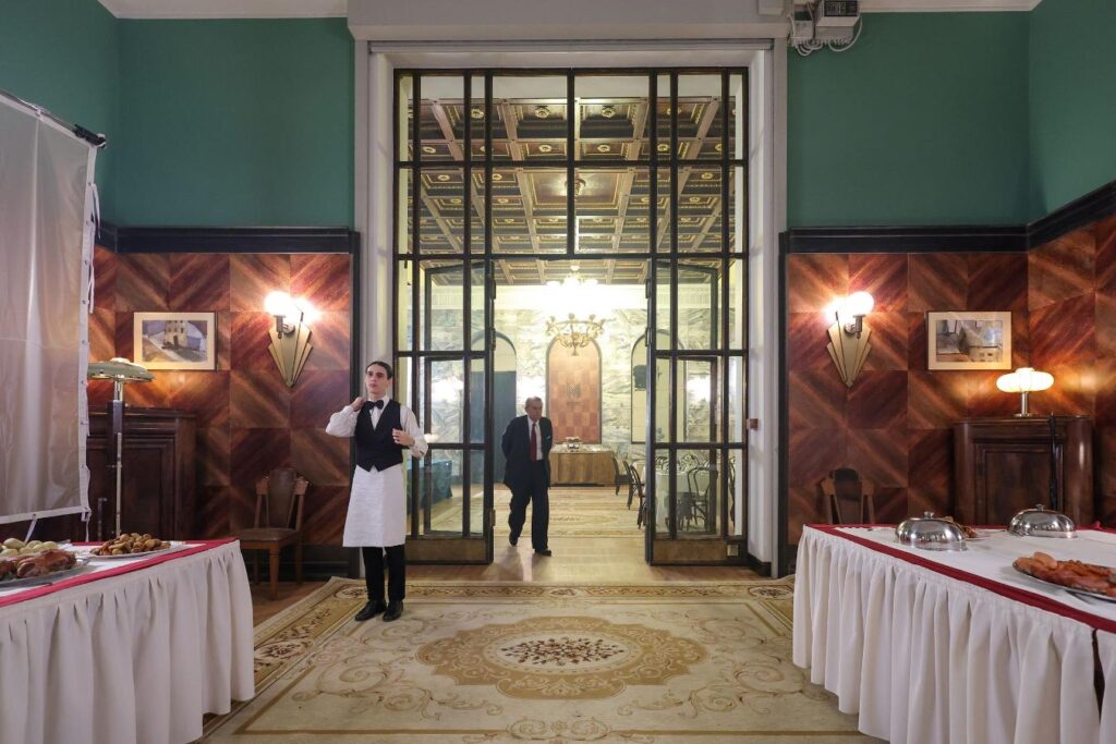 Kelner w eleganckim stroju poprawia muszkę, stojąc przy stołach z jedzeniem w stylowym wnętrzu restauracji.