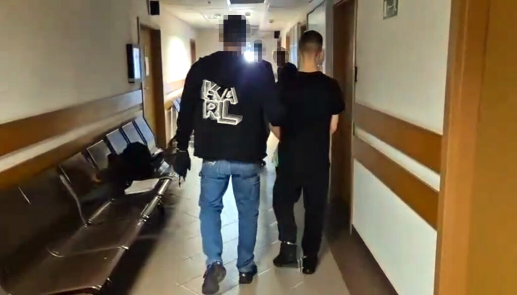 Mężczyzna w czarnej kurtce prowadzi drugiego mężczyznę w czarnym ubraniu przez szpitalny korytarz.