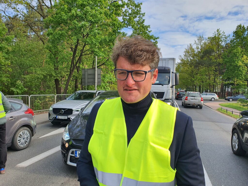 Mężczyzna w kamizelce odblaskowej stoi obok ruchliwej ulicy, z samochodami w tle i zielonymi drzewami.