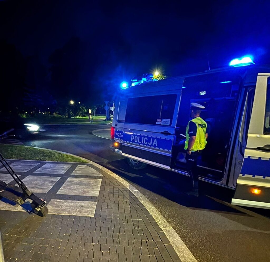 Policjant w kamizelce odblaskowej stoi obok radiowozu z włączonymi sygnałami świetlnymi na skrzyżowaniu w nocy.