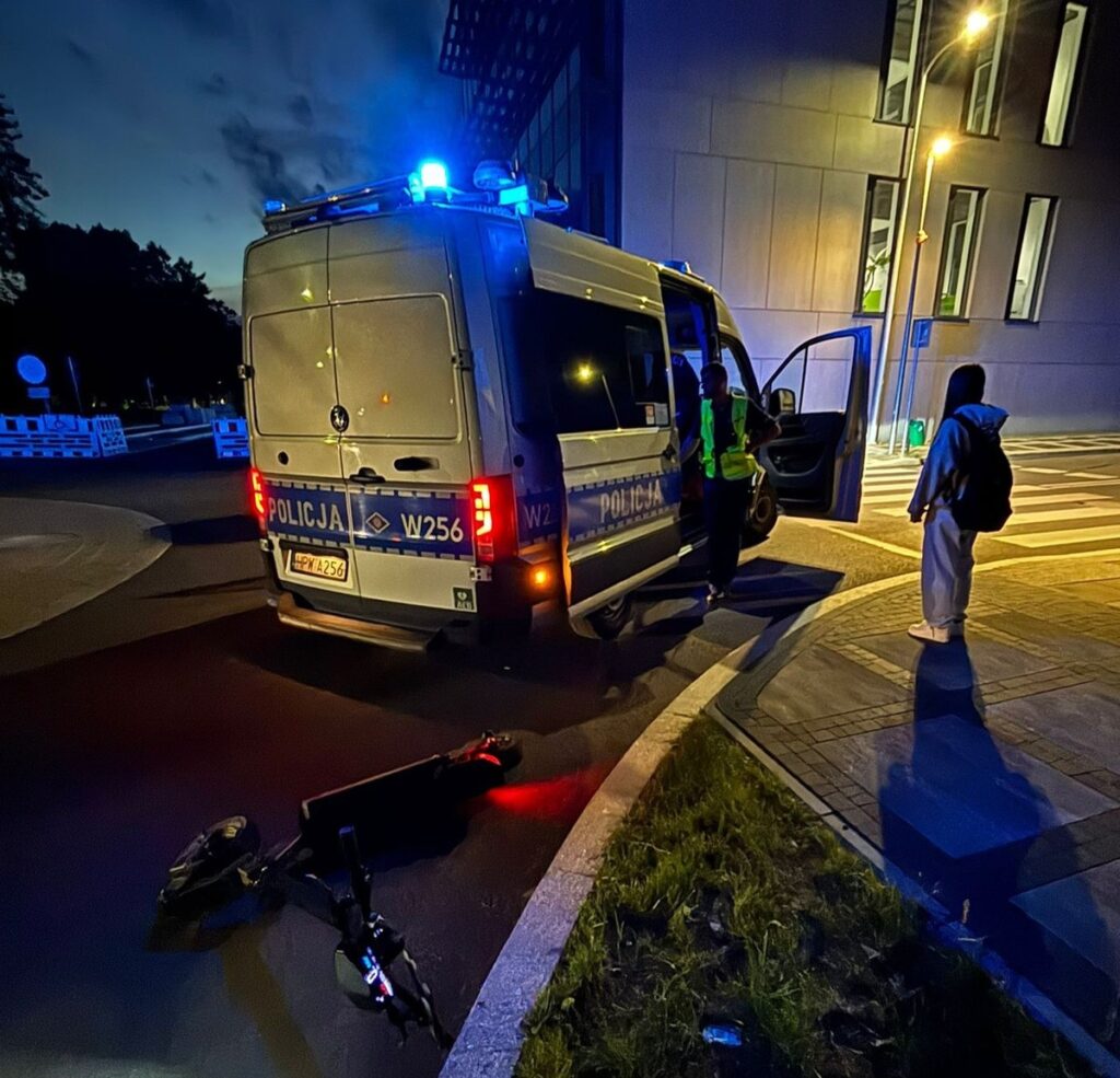 Policjant rozmawia z osobą stojącą obok przewróconej hulajnogi w miejskim otoczeniu nocą.