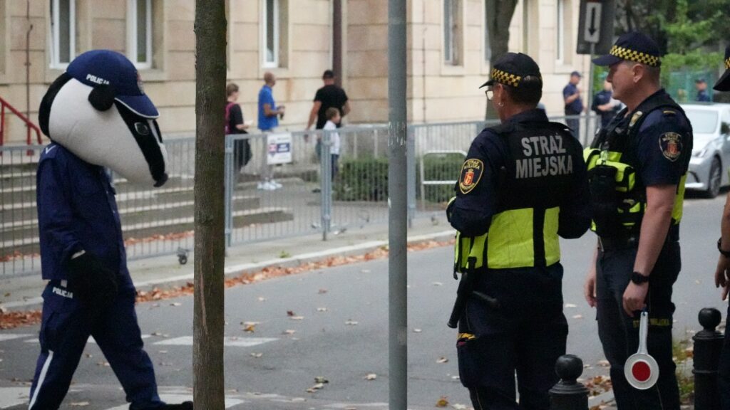 Mężczyzna w kostiumie maskotki z głową psa spacerujący obok funkcjonariuszy straży miejskiej na ulicy.