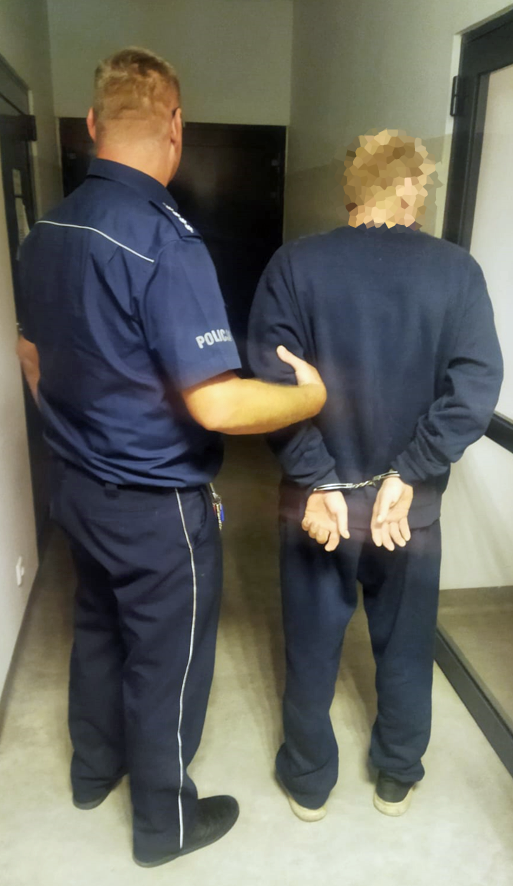 Policjant prowadzi zatrzymanego mężczyznę w kajdankach w korytarzu budynku.