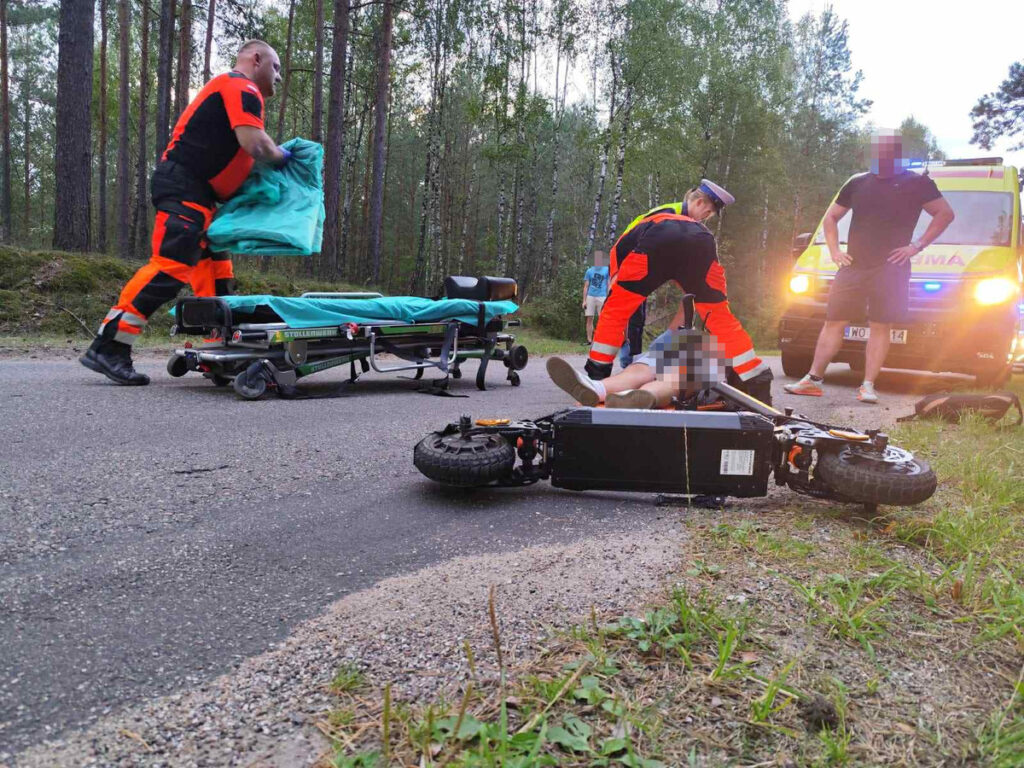 Ratownicy medyczni udzielają pomocy poszkodowanemu na drodze w lesie, obok przewróconego skutera.