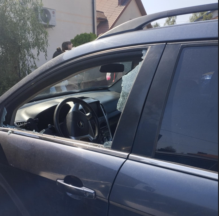 Uszkodzone auto z wybitym oknem, w środku widoczna kierownica, w tle osoby stojące obok budynku.