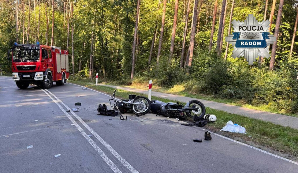 W pobliżu drogi leży przewrócony motocykl, a obok niego rozrzucone elementy odzieży i kask. W tle widoczny jest wóz strażacki.