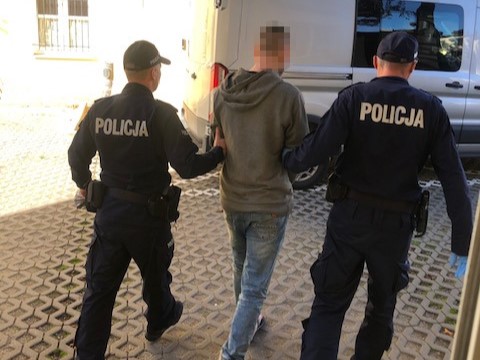 Dwóch policjantów prowadzi zatrzymanego mężczyznę w szarej bluzie na podwórzu.