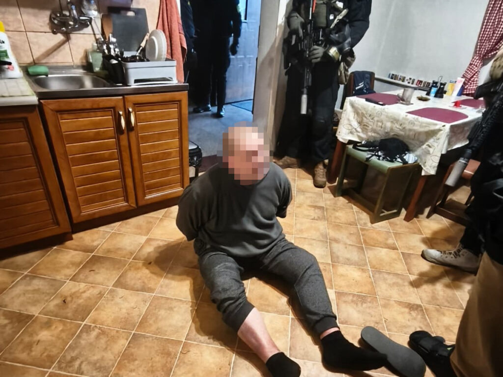Mężczyzna w szarej odzieży siedzi na podłodze w kuchni, z rękami związanymi z tyłu, podczas akcji służb mundurowych.