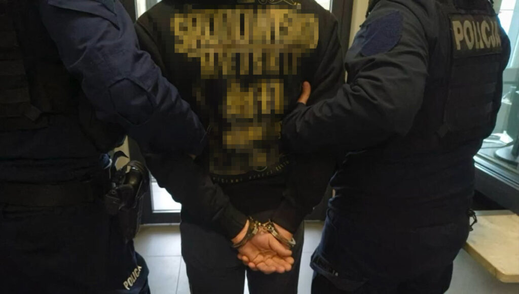 Mężczyzna w kajdankach zatrzymywany przez dwóch policjantów w pomieszczeniu z dużymi oknami.