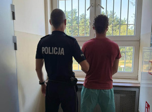 Policjant w mundurze stoi za mężczyzną w koszulce, obaj patrzą przez okno w pomieszczeniu z kratami.