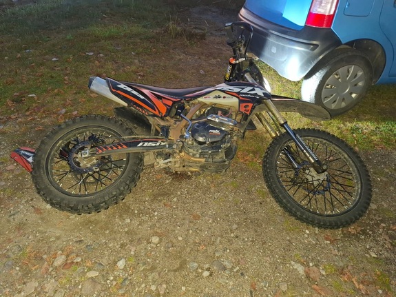 Motocykl terenowy o pomarańczowo-czarnym malowaniu stoi na ziemnym podłożu obok niebieskiego samochodu.