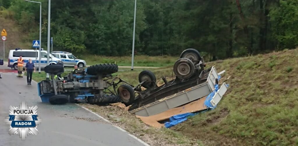 Traktor przewrócony na bok obok drogi, z grupą ludzi oraz radiowozem policji w tle.