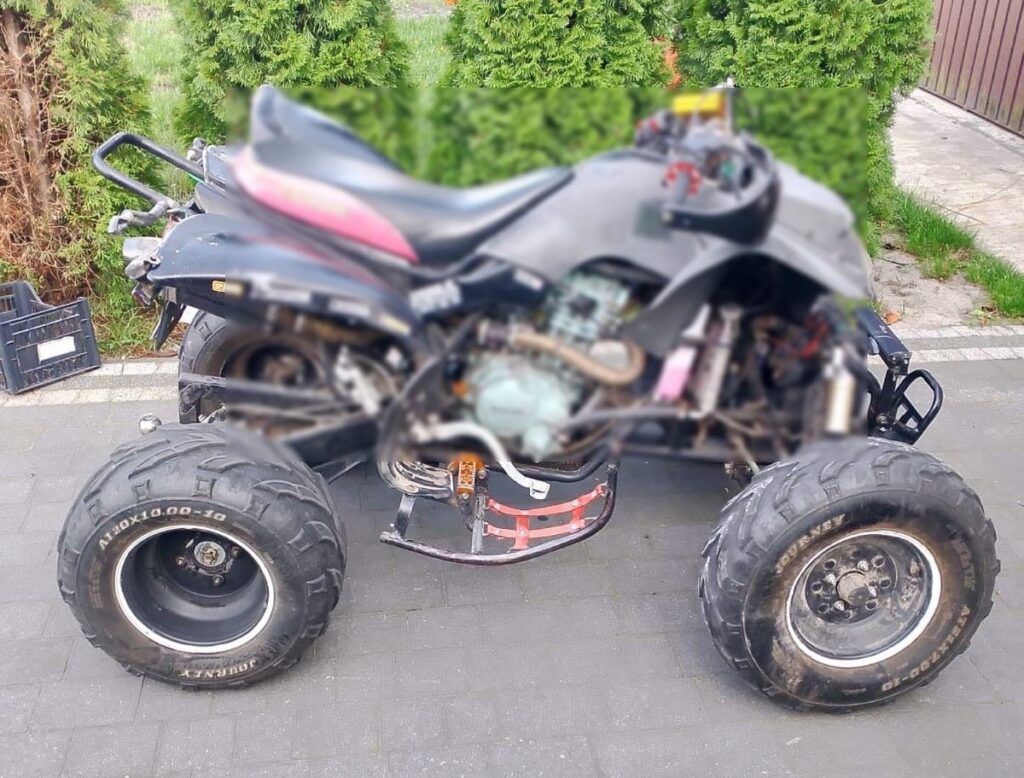 Czarny quad z dużymi oponami stoi na kostce brukowej, w tle widoczne krzewy i ogrodzenie.
