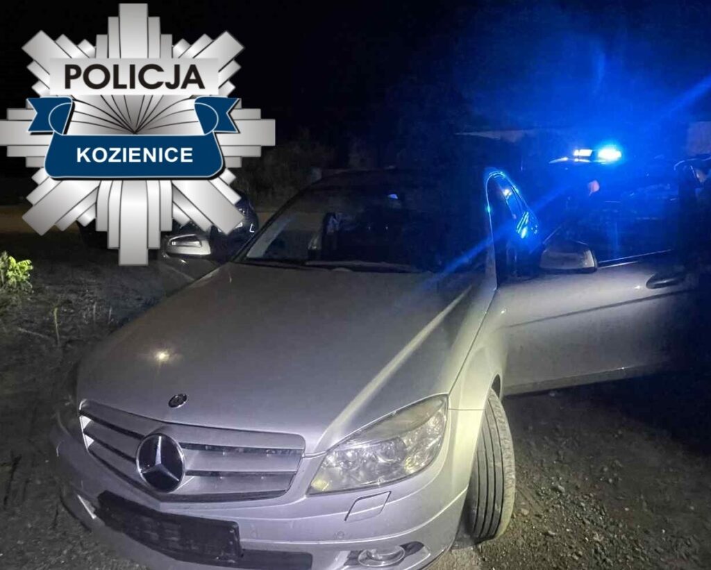 Policyjny radiowóz z niebieskimi światłami stoi obok srebrnego samochodu osobowego zaparkowanego na nieutwardzonej drodze.
