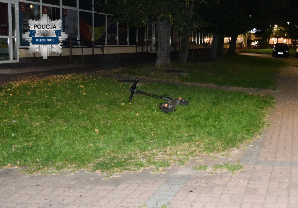 Na trawie leży elektryczna hulajnoga w pobliżu oświetlonego chodnika i budynku.