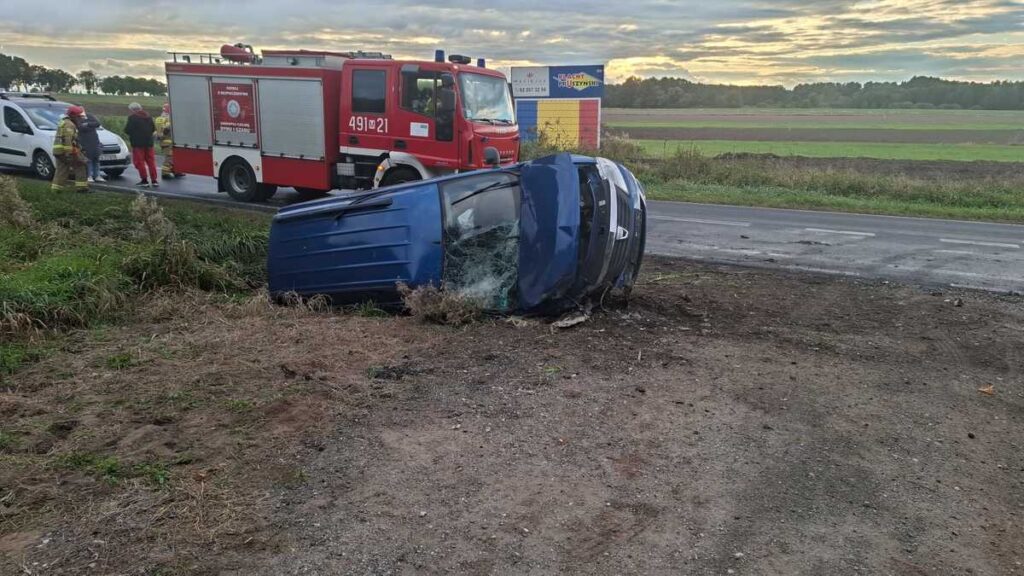 Nieoznakowany niebieski samochód leży na boku przy drodze, obok stoi wóz strażacki oraz kilka osób w mundurach.