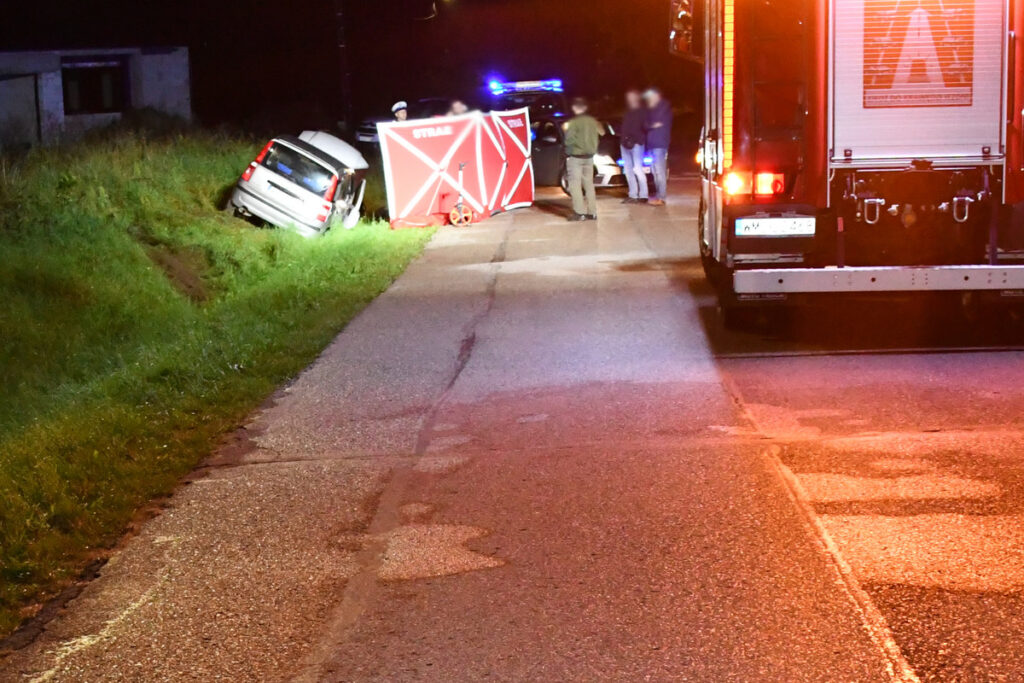 W nocy na drodze leży przewrócone auto osobowe, obok grupka osób z policji i straży pożarnej.
