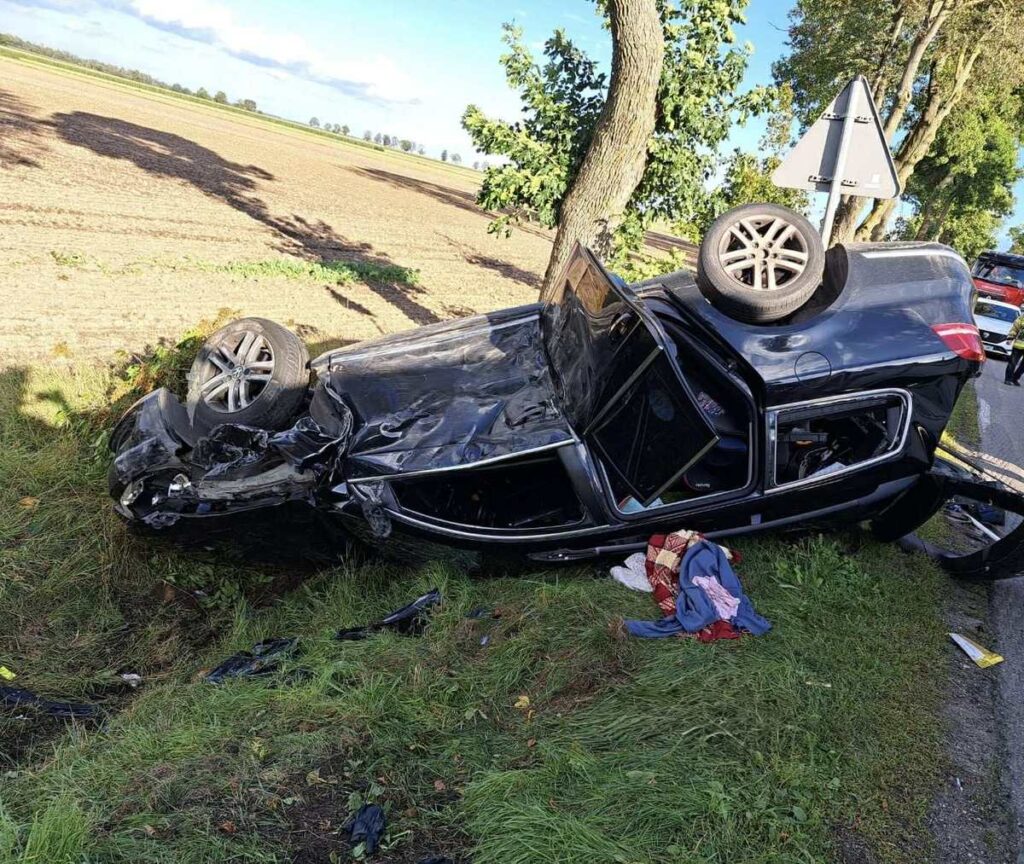 Czarny samochód leży na dachu w rowie, obok drogi, z uszkodzoną karoserią i porozrzucanymi ubraniami na trawie.