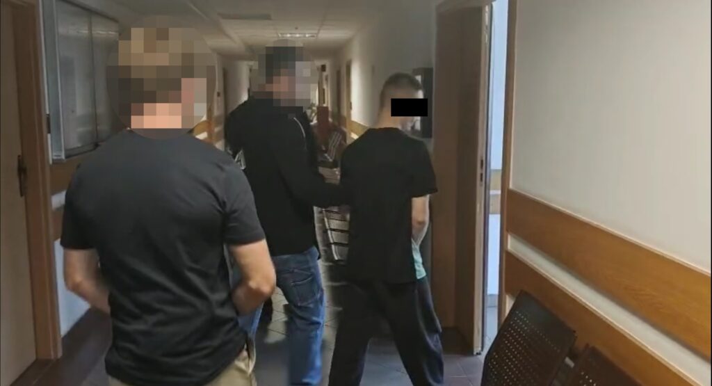 Mężczyzna w czarnej bluzie prowadzi młodego mężczyznę w kierunku drzwi w korytarzu budynku.