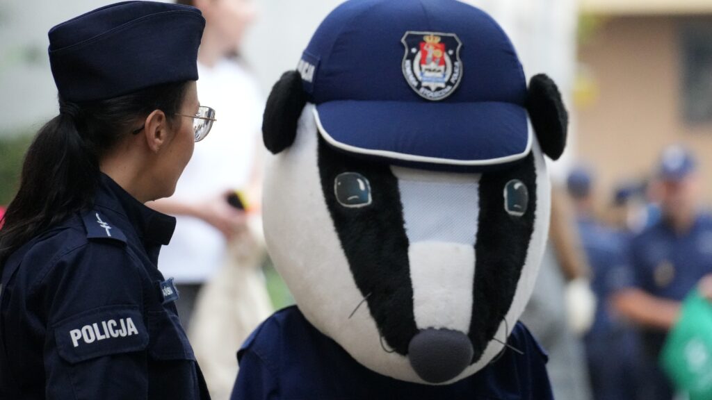 Policjantka rozmawia z osobą przebranym za maskotkę jeża w mundurze policyjnym podczas wydarzenia na świeżym powietrzu.