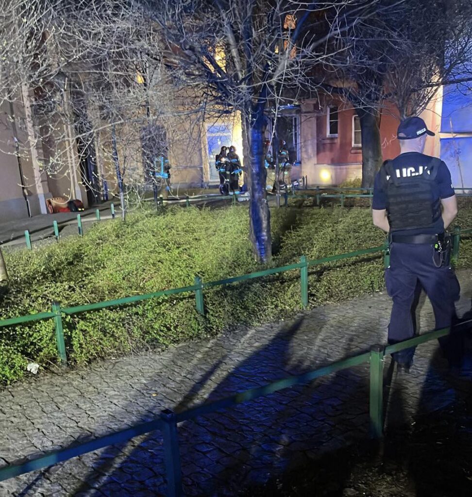 Policjant w kamizelce ochronnej obserwuje akcję ratunkową, w której biorą udział strażacy w pobliżu budynku.